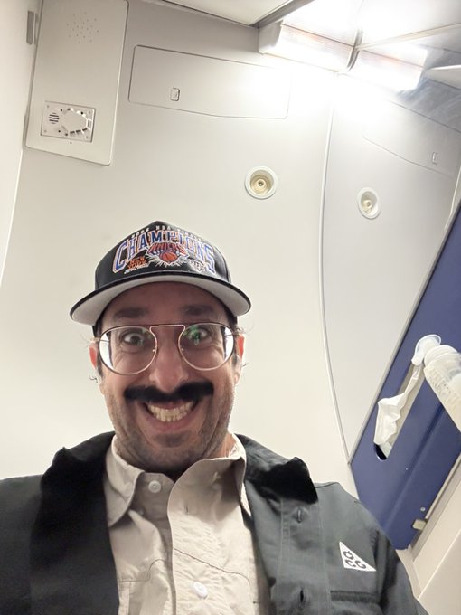 A man with glasses and a mustache wearing a 'Champions' baseball cap takes a selfie inside what appears to be an airplane lavatory.