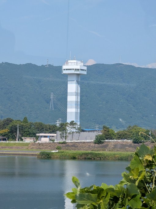 A scenic photo of a tall observation tower beside a calm lake with green mountains and power transmission towers in the background.
