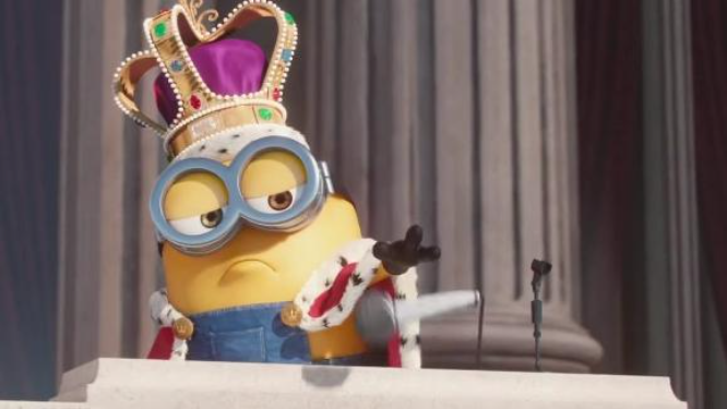 A Minion character wearing a jeweled crown and glasses, dressed as a king, standing at a podium with a microphone and gesturing with a gloved hand.