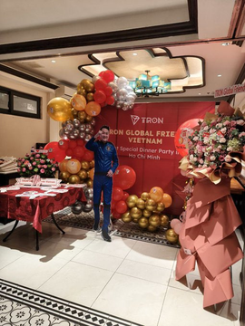 The image features a man standing next to a large balloon arch that says "TRON Global Free Vietnam" above him. The balloon arch is made up of red, gold, and silver balloons arranged in an impressive display. In front of the balloon arch, there are tables with food and drinks set up for guests attending a special dinner party. A man can be seen standing next to one of these tables, possibly enjoying the event or waiting to join others at the gathering.