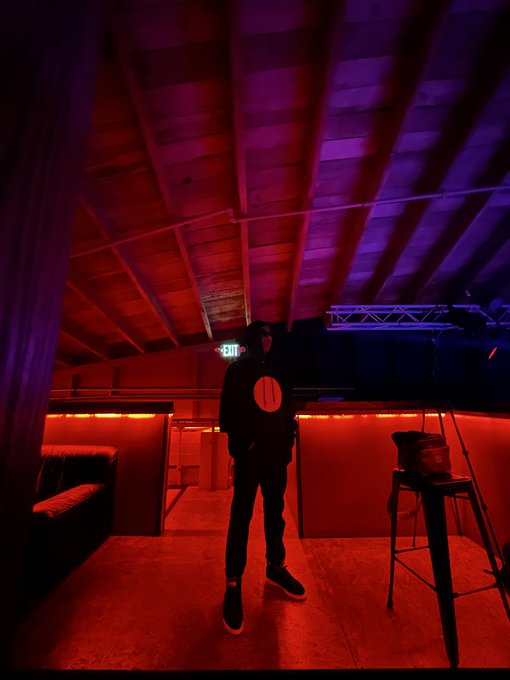 A person wearing a black hoodie with a red circular logo stands in a dimly lit room illuminated by red and purple lights, with a wooden ceiling and bar stools visible.