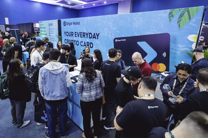 A crowded convention hall where attendees gather around a bright blue Bitget Wallet booth promoting "YOUR EVERYDAY CRYPTO CARD."