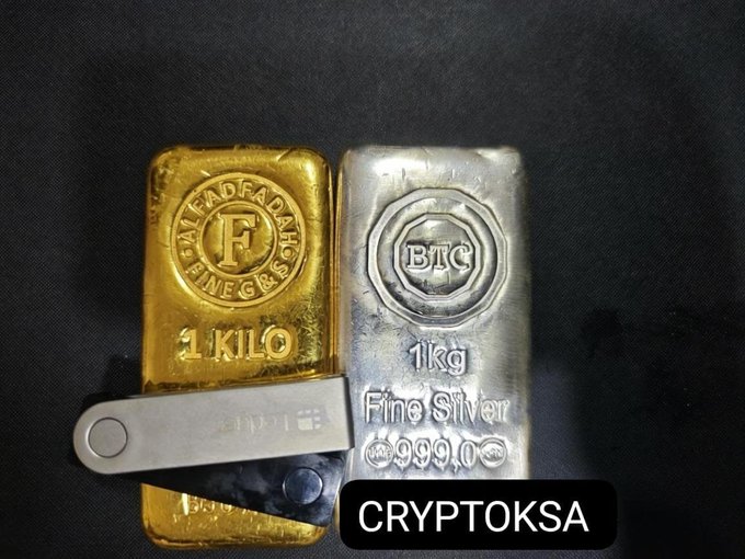 A 1 kilo gold bar stamped "ADFAD FINE GR" and a 1kg BTC "Fine Silver" 999.0 bar rest on black fabric alongside a Ledger hardware wallet, visually linking traditional precious metals to cryptocurrency storage, as highlighted by the "CRYPTOKSA" caption.