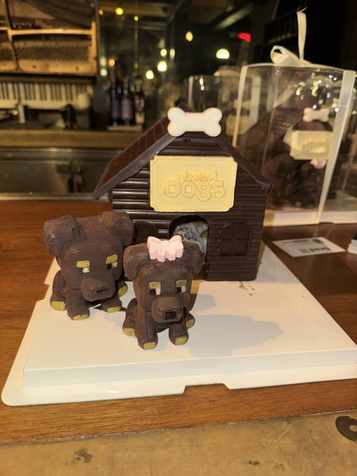 A chocolate sculpture of two dogs, one wearing a pink bow, sitting in front of a dog house labeled 'Hotel dogs' with a bone decoration on the roof.