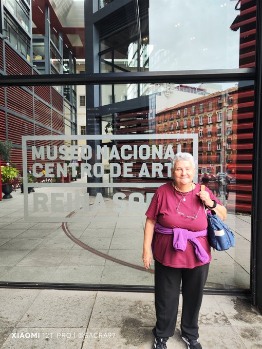 An elderly woman in a pink shirt and purple sweater tied around her waist smiles in front of the glass entrance of the **Museo Nacional Centro de Arte Reina Sofía** (with "REINA SOFÍA" partially visible), capturing her visit to the famous Spanish art museum.