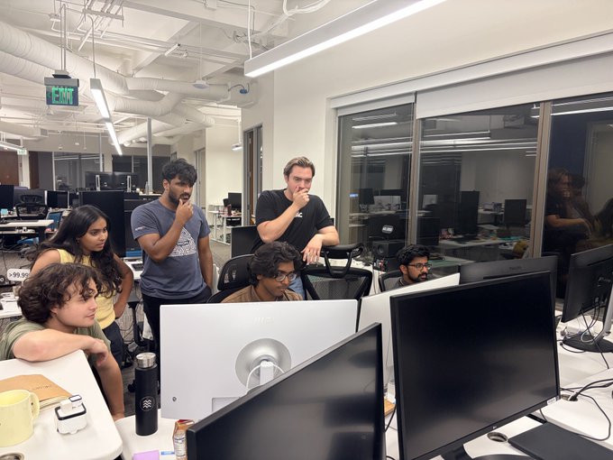 A team at Matic Robots is gathered around a monitor in an open office, appearing engaged in a collaborative discussion following a major breakthrough, which they will showcase on 8/13.