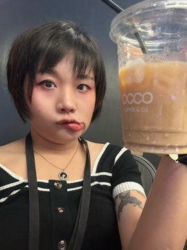 A selfie of a young woman with short dark hair wearing a black and white striped top, playfully holding a clear plastic cup of iced coffee from the brand 'COCO'.