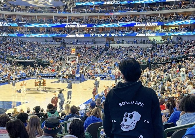 A person wearing a black 'BORED APE' hoodie stands in the foreground of a packed basketball arena during what appears to be a Kansas vs. Kentucky game.