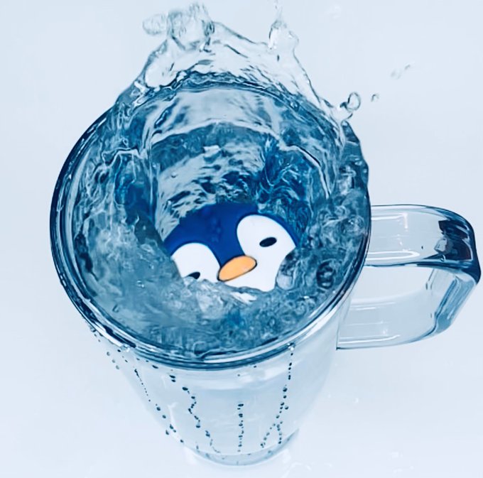 A clear blue glass mug splashes with water, revealing a cartoon penguin face at the bottom of the cup.