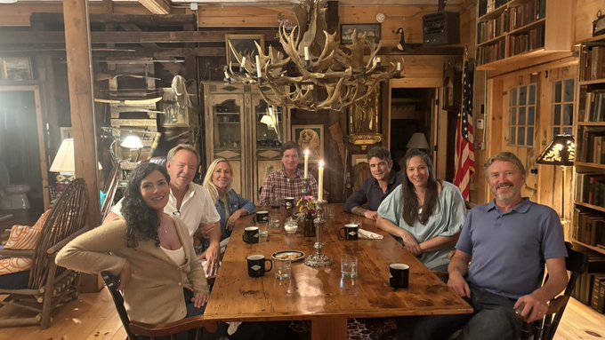 A group of seven people posing around a long wooden dining table in a rustic room featuring an antler chandelier, an American flag, and bookshelves.