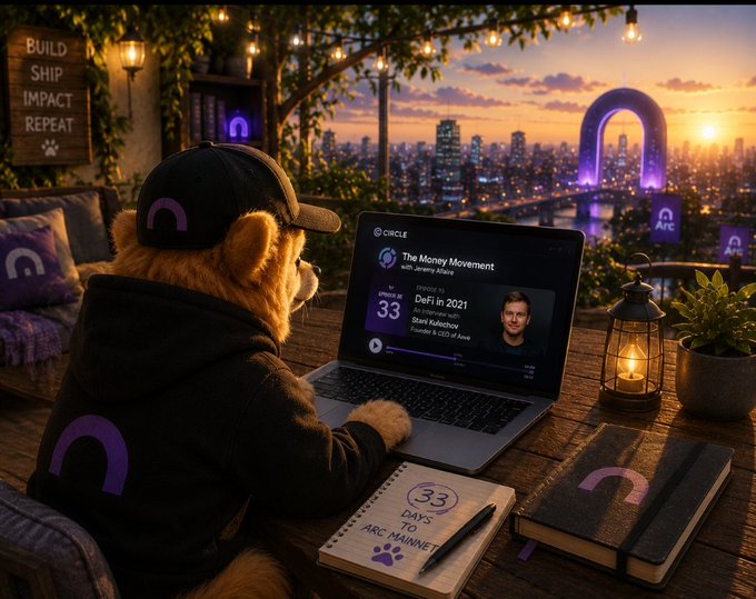 An anthropomorphic teddy bear wearing a black cap and hoodie sits at a rooftop desk with a city skyline at sunset, watching Episode #33 of "The Money Movement" on a laptop featuring Stani Kulechov (Aave), while a notebook marked "33 Days to Arc Mainnet" reinforces the countdown theme.