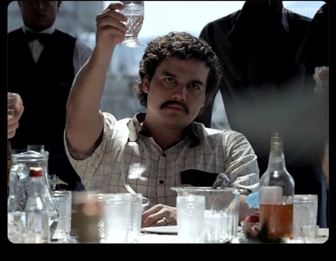 A scene from a film showing a man with a mustache in a checkered shirt raising a glass in a toast, with waiters standing behind him and a table full of bottles and glasses in the foreground.