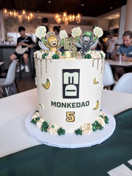 A white birthday cake decorated with bananas and 'MONKEDAO 5' branding, topped with two cartoon monkey figures wearing crypto-related accessories.