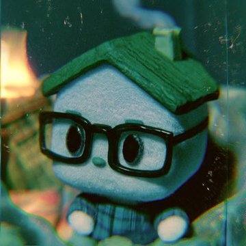 A small felt craft figure with a green roof-like hat, large black glasses, and a plaid shirt is displayed behind glass.
