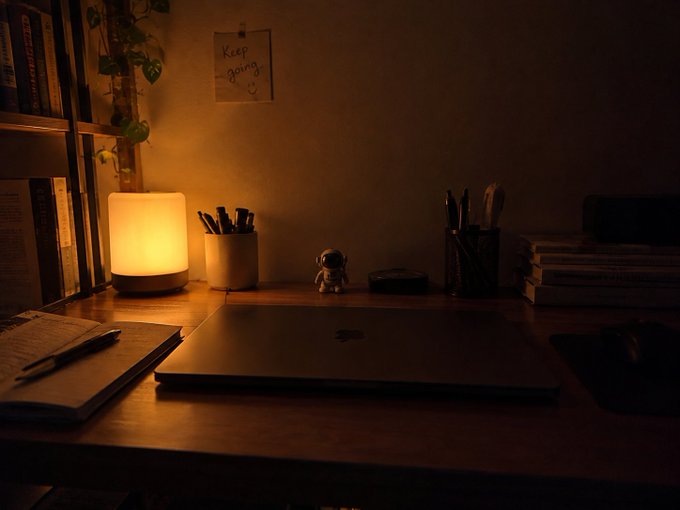 A dimly lit desk setup features a warm glowing cylindrical lamp, a closed laptop, an open notebook, pen holders, a small astronaut figurine, and a 'Keep going' note pinned to the wall.