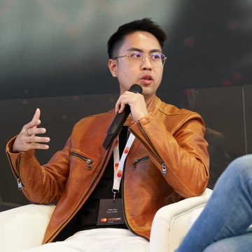 A young man wearing glasses and a brown leather jacket speaks into a microphone while gesturing with his hand, seated at what appears to be a panel or interview setting. His name badge and lanyard are visible, though the text is not clearly legible, and his engaged expression aligns with the post's context of discussing Trust Wallet's priorities regarding transaction speed and slippage.