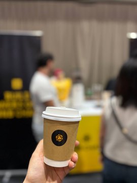 A hand holds up a brown coffee cup with a white lid featuring a small logo, set against a blurred background of a convention or expo hall with yellow branding visible.