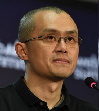 A close-up photograph of a bald man wearing glasses and a black shirt, appearing to speak into a microphone.