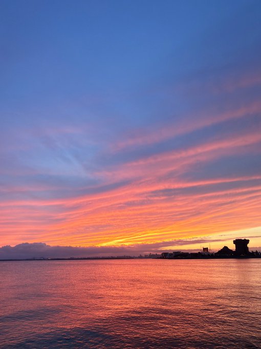 A dramatic sunset over a body of water features streaks of pink and orange clouds, with a dark silhouette of a distant shoreline and structures.