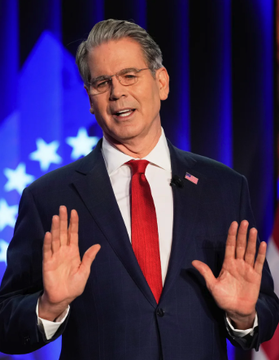 A man wearing glasses, a dark blue suit, and a red tie stands on a stage with both hands raised, with an American flag visible in the blue-lit background.