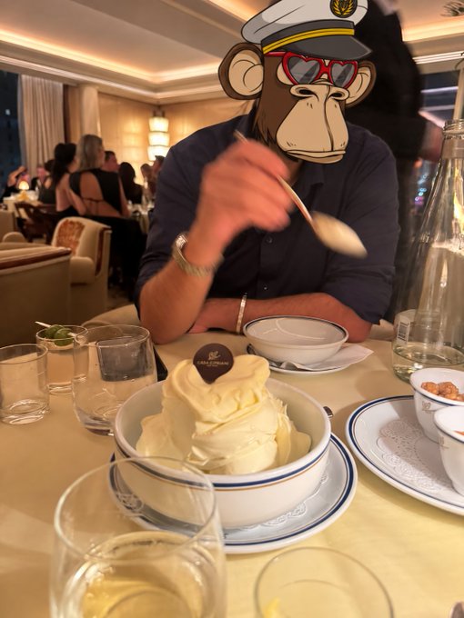 A person in a dark shirt sits at a restaurant table with a large dish of soft-serve ice cream, their face obscured by a cartoon monkey sticker wearing a captain's hat and heart-shaped sunglasses.