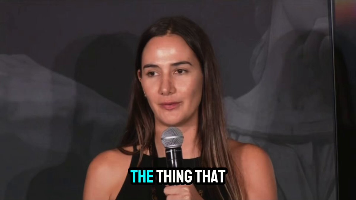 The image features a woman standing next to a microphone while speaking into it. She appears to be delivering a speech or presentation, as indicated by her body language and the presence of the microphone. The background of the image is dark gray, which contrasts with the woman's figure and makes her stand out more prominently.