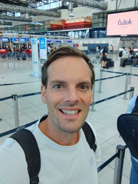 A selfie of a smiling man wearing a backpack at an airport terminal, with a store sign reading 'Cede' visible in the background.