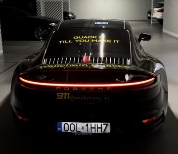 A black Porsche 911 Carrera 4S is shown from the rear in a parking garage, displaying yellow text on its rear window that reads "QUACK TILL YOU MAKE IT" and "WALLCHAIN QUACKS," along with the contact info "911@quackiverse_4s" and a license plate reading "OOL 1H7." The image relates to the post by showcasing the luxury sports car reportedly purchased with Wallchain Quacks, highlighting the founder's success.