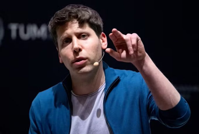 A man with short brown hair, wearing a blue jacket over a gray shirt and a headset microphone, is captured mid-speech while gesturing with his right hand raised, with partial text "TUM" visible in the background. The image appears to be of Sam Altman, an American tech founder, connecting to the post's theme comparing him to Do Kwon, the Korean crypto founder behind the $LUNC token.