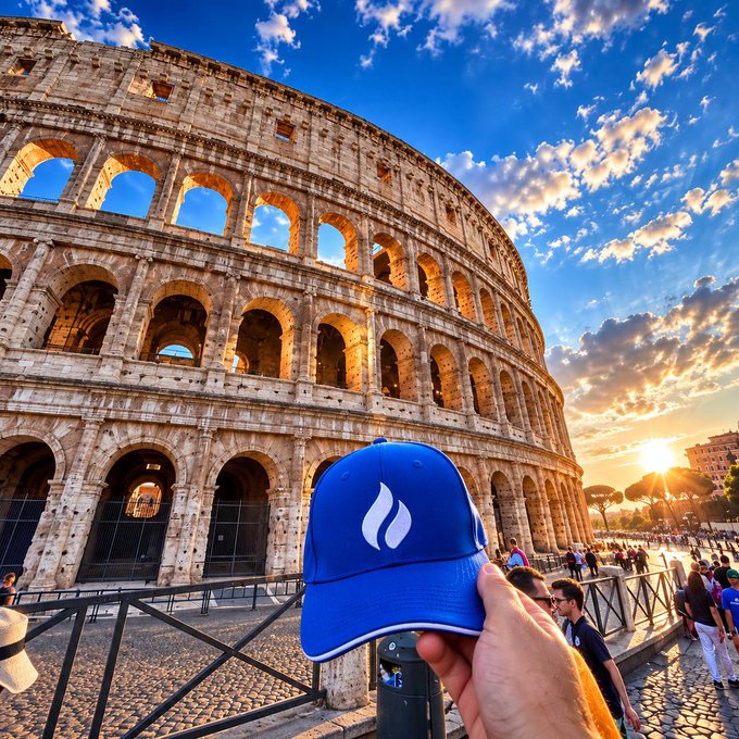 A hand holds a blue cap featuring the "HTX" logo in front of the iconic Colosseum in Rome at sunset, visually linking the ancient marvel's enduring construction to HTX's ongoing growth and development. The image cleverly draws a parallel between Rome's timeless legacy and the steady building of a modern enterprise.