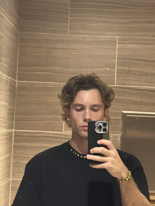 A young man with wavy hair takes a mirror selfie in a tiled bathroom, wearing a black t-shirt with partially visible text 'AJOMUD', a gold chain, and a gold bracelet.