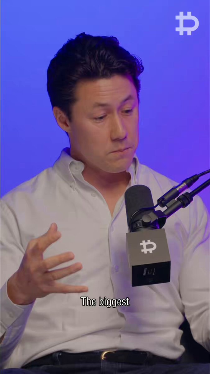 The image features a man wearing a white shirt, sitting down with his hands outstretched as if he's explaining something or making a gesture. The background of the image has a blue hue, which contrasts with the man's attire. There are also some words visible in the image that seem to be related to Bitcoin or cryptocurrency.