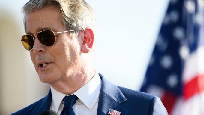 A man in a blue suit wearing aviator sunglasses speaks into a microphone, with an American flag visible in the background.