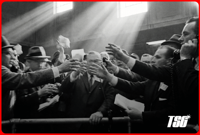 A vintage black-and-white photograph of a busy stock trading floor, showing men in suits shouting and reaching out with paper orders under dramatic beams of light, with a 'TSG' watermark.