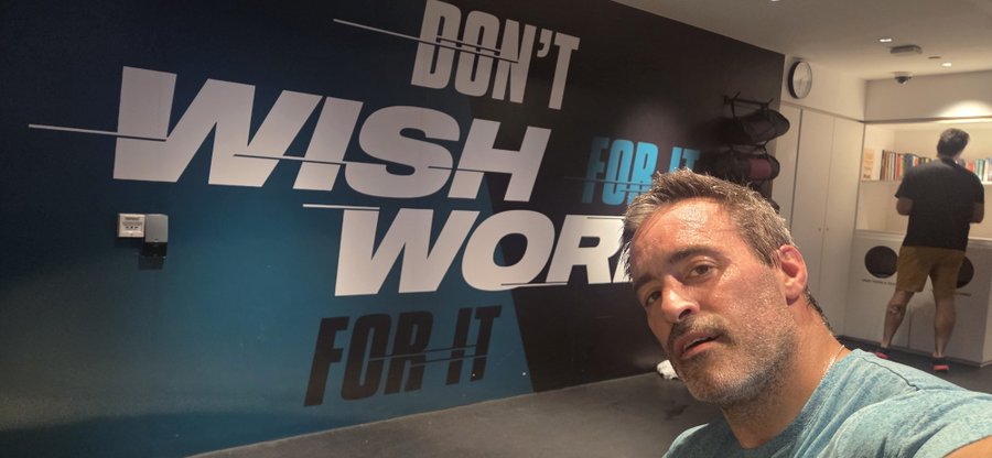 A man takes a selfie in front of a motivational gym wall that reads "DON'T WISH FOR IT, WORK FOR IT," with another person working out near exercise equipment in the background. The bold wall text perfectly reinforces the post's message, reminding viewers that achieving fitness goals requires effort rather than mere desire.