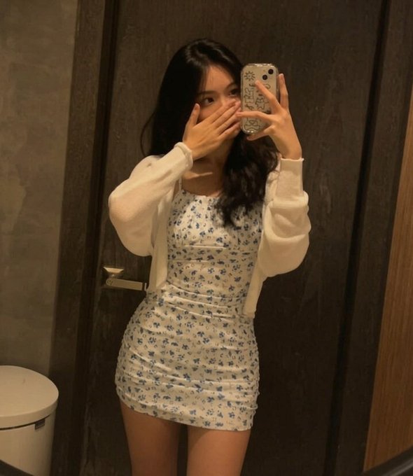 A mirror selfie of a young woman with long dark hair covering part of her face, wearing a white floral mini dress under a white cardigan in what appears to be a bathroom.