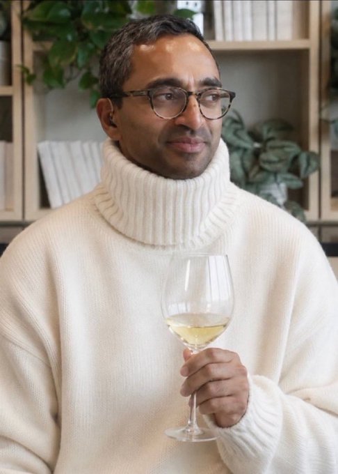 A man wearing glasses and a cream turtleneck sweater holds a glass of white wine, standing in front of a bookshelf with plants.