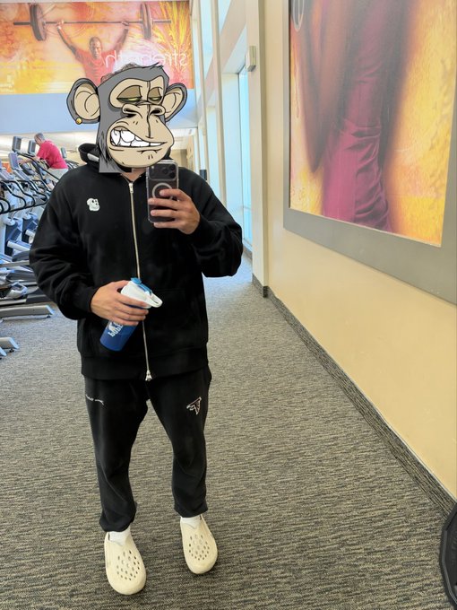 A person in a black tracksuit and white crocs takes a mirror selfie in a gym, with a cartoon monkey sticker covering their face.