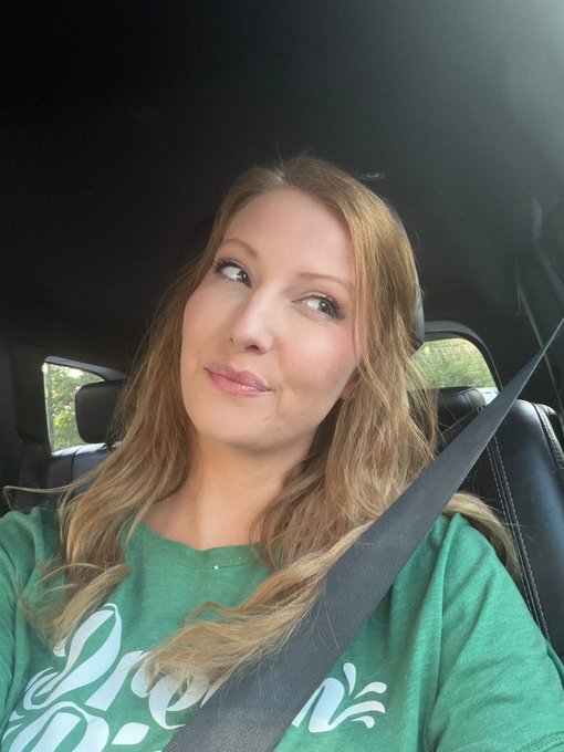 A woman with long blonde hair wearing a green graphic t-shirt sits in the driver's seat of a car, secured by her seatbelt, with a slight smile as she glances upward. The casual selfie complements the "gm future @UseArtisan users" post, suggesting a friendly morning greeting directed at the community.