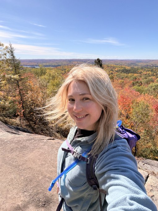 A blonde woman takes a selfie while hiking on a rocky overlook, with a vibrant autumn forest of red, orange, and green foliage stretching into the distance behind her.