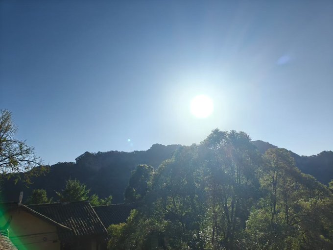 A bright sun shining in a clear blue sky above a tree-covered mountain ridge with traditional rooftops visible in the foreground.
