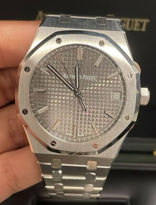 The image features a silver watch with a square face, which appears to be an Aquafina brand watch. The person holding the watch has their hand visible above the wristwatch, showcasing its design and craftsmanship. The background of the image is black, making the watch stand out even more. There are no other objects or texts in the image, so it focuses solely on the watch and the person's interaction with it.