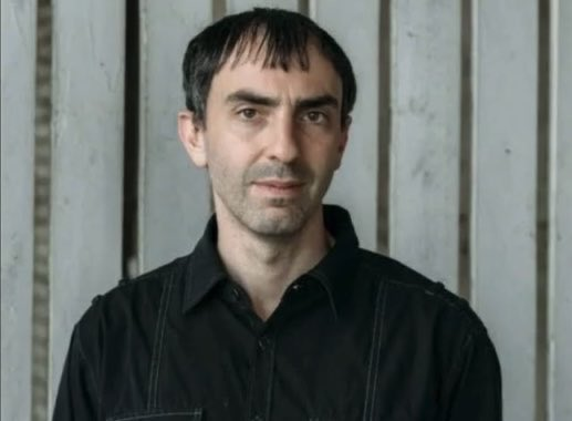 A man with dark hair and a black shirt stands against a corrugated metal wall, his expression serious and contemplative—fitting for someone portrayed as opposed to the BIP-110 Bitcoin proposal.