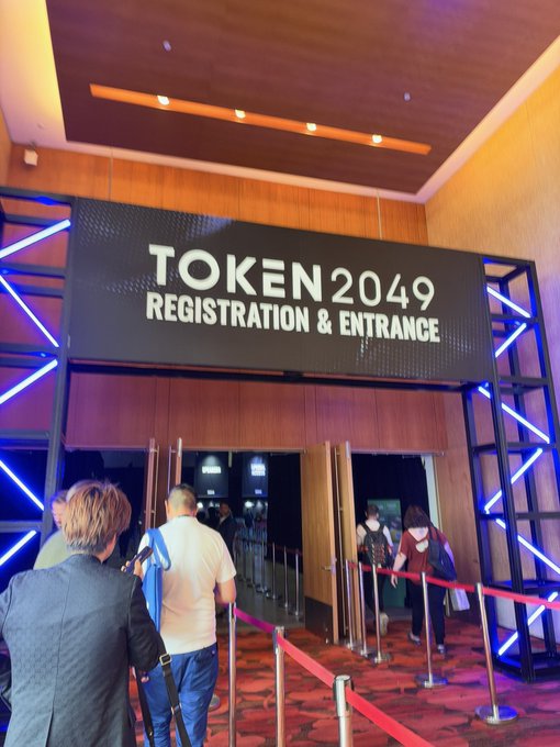 People queue at the entrance of a conference beneath a sign reading "TOKEN 2049 REGISTRATION & ENTRANCE", framed by blue neon lights.