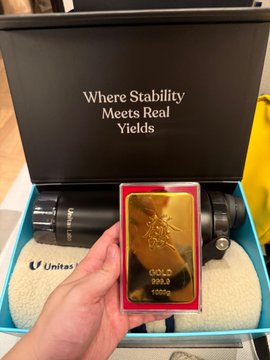 A hand holds up a gold bar in front of an open black presentation box that reads 'Where Stability Meets Real Yields', with a camera lens and 'Unitas' branded case visible inside the packaging.