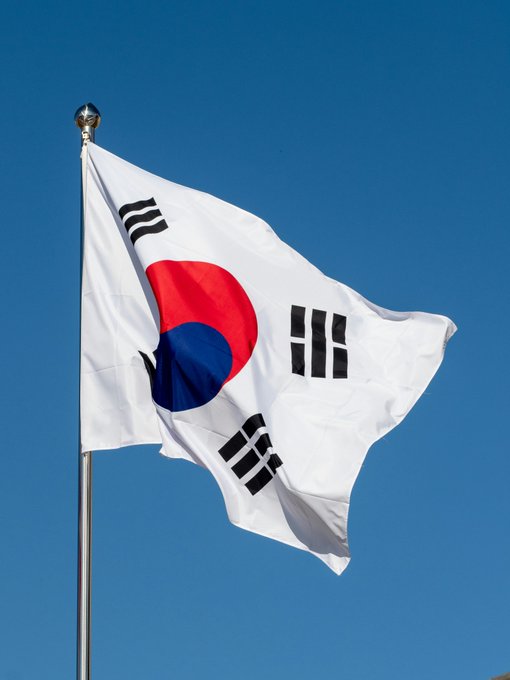 A photograph of the South Korean national flag (Taegukgi) waving on a flagpole against a clear blue sky, displaying the red and blue taegeuk symbol with four black trigrams.