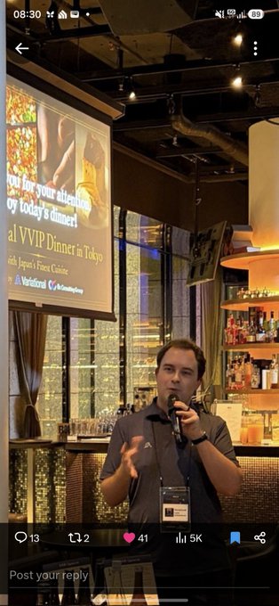A man speaks into a microphone at what appears to be a Variational.io "VVIP Dinner in Tokyo with Aspen's Finest Cuisine," as shown on the bar's illuminated sign behind him, fulfilling the poster's playful aspiration to attend the exclusive event.