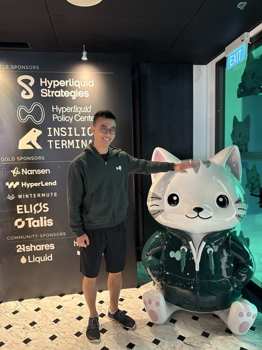A photograph of a smiling young man wearing glasses and a dark green hoodie, posing with his hand on a large cat mascot statue at an event with sponsor logos including Hyperliquid Strategies, Insilico Terminal, Nansen, HyperLend, Wintermute, EliΞS, Talis, 21shares, and Liquid visible on the wall behind him.