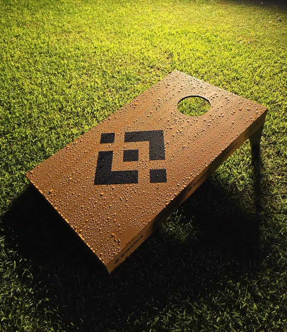 A wooden cornhole board sitting on green grass with water droplets and a black geometric logo on its surface.