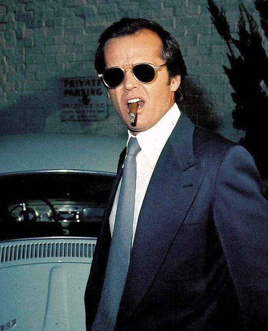 The image features a man dressed in a suit, wearing sunglasses and holding a cigar in his mouth. The man appears to be smoking the cigar while standing next to an old car. There are also other cars visible in the background of the image. The caption provided seems to be unrelated or unrelated to the main subject matter of the photo.